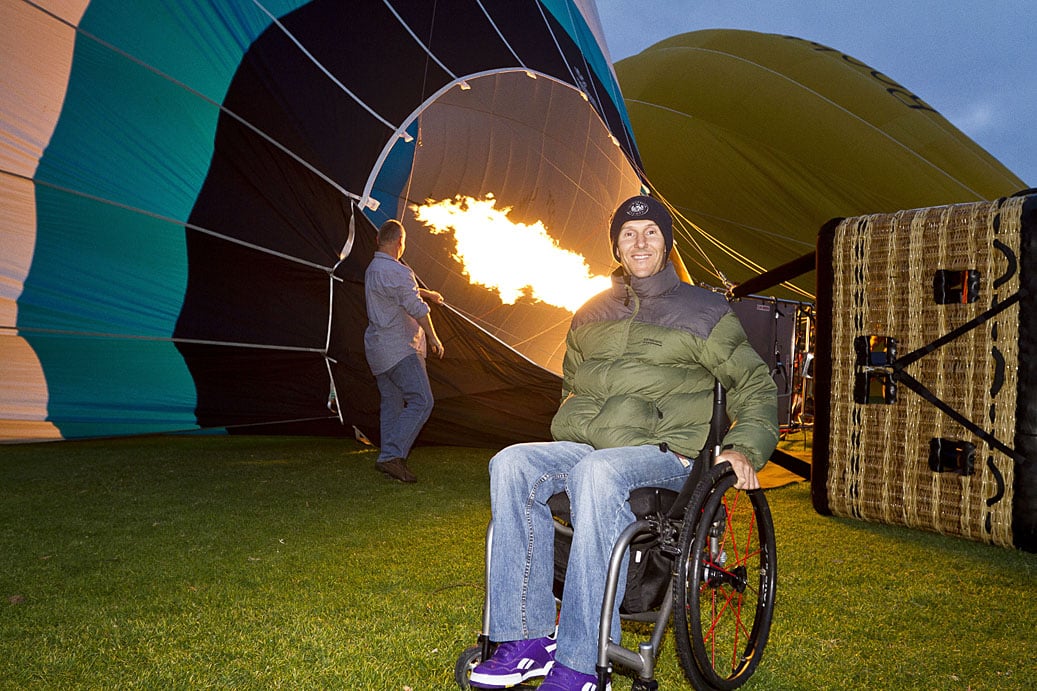 Smiling man in a wheelchair next to a hot air balloon being inflated with a large flame.