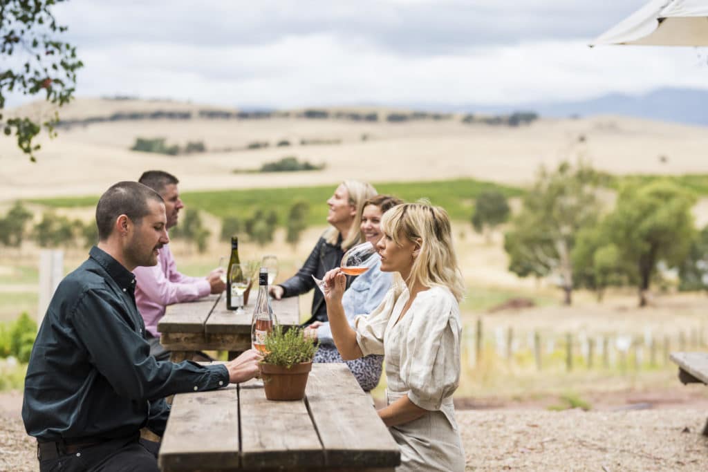 A group of friends enjoying a wine at timber tables outside with vineyard and mounrtain views at Delatite Winery