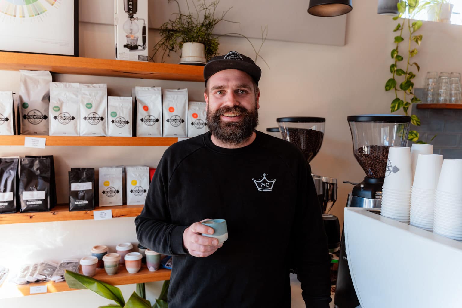 Luke, a male cafe owner and coffee roaster, stands with a ceramic mug in front of a coffee machine.
