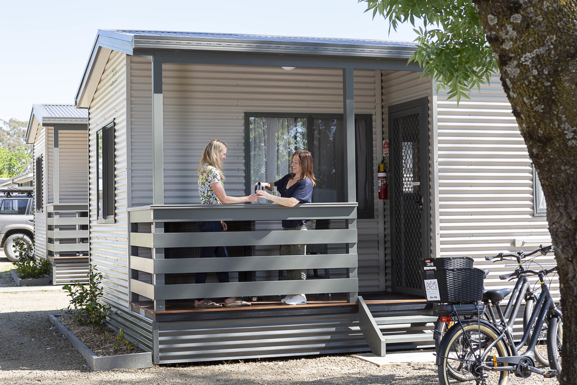 Two people relaxing with a glass of wine on the veranda of a cabin at Valley View Caravan Park with bike in the background in Whitfield