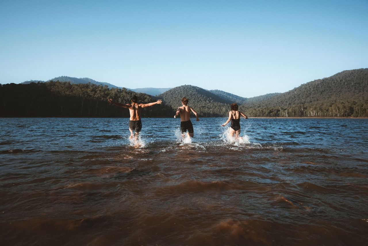 Three people running into the waters of Lake William Hovell with mountains in the background