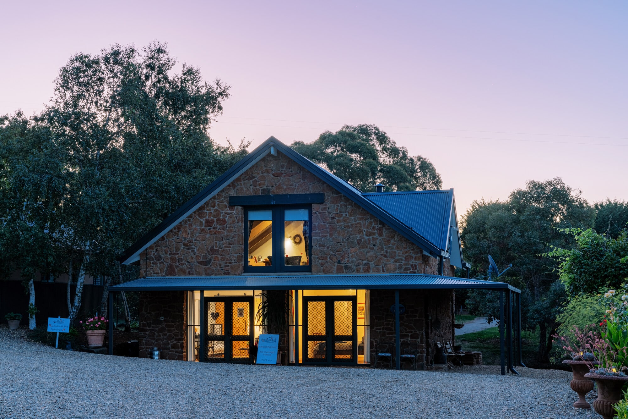 A rustic stone building at dusk, with lights on inside and trees and flower pots surrounding it.