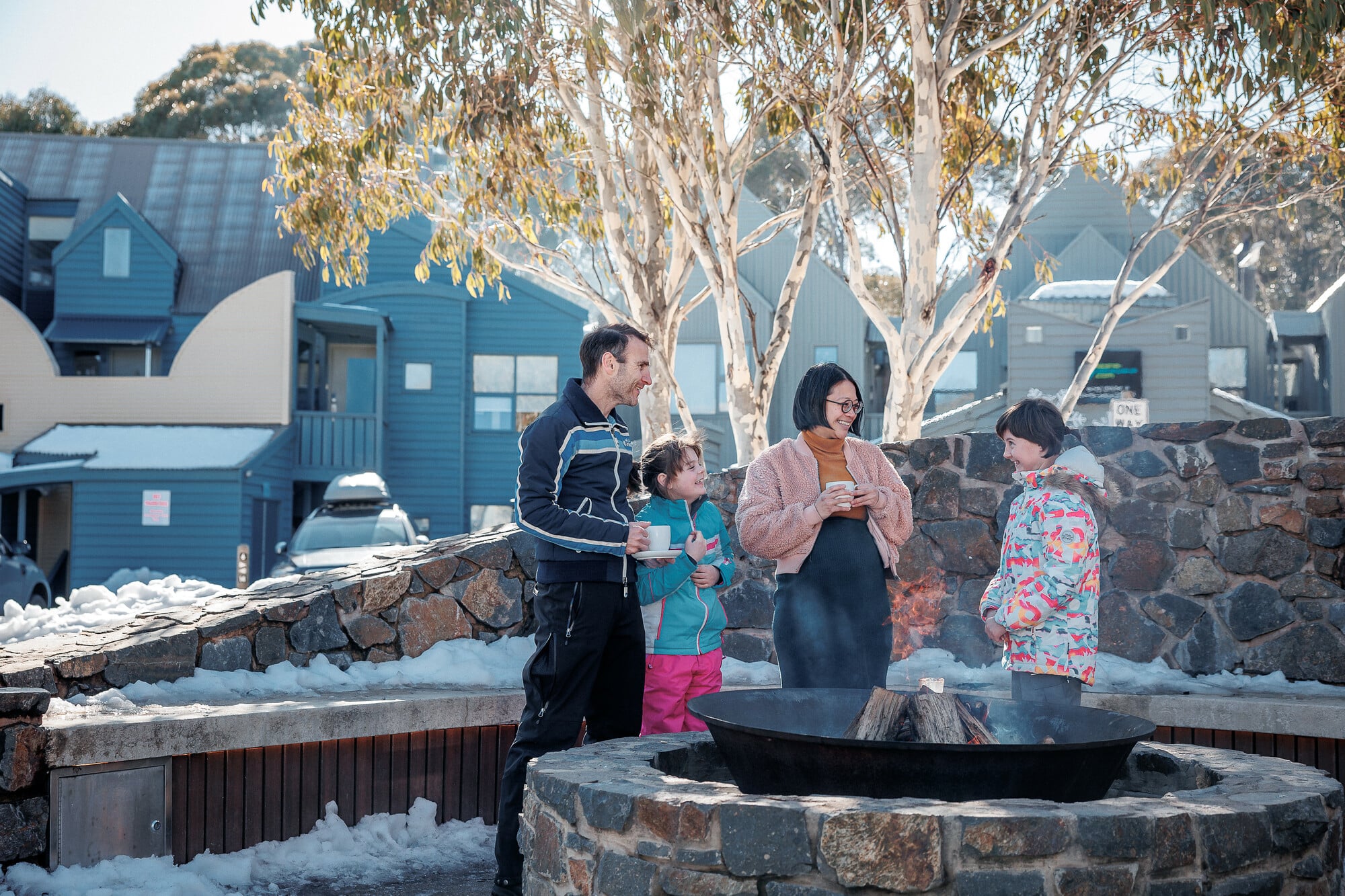 Two adults and two children sipping drinks and talking around the fire pit at Dinner Plain with snow in the background