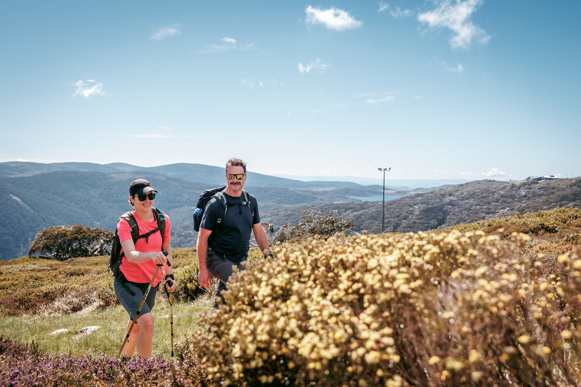 Two people walking through wildflower meadows on a hiking trail in Falls Creek with mountain views and a lake in the background.