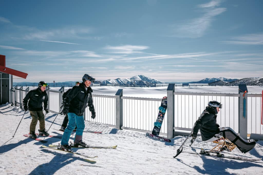 Adaptive skier with Disabled Wintersport Australia guides skiing from the Alpine Gateway Hub at Mt Hotham with snowy mountain views in the background.