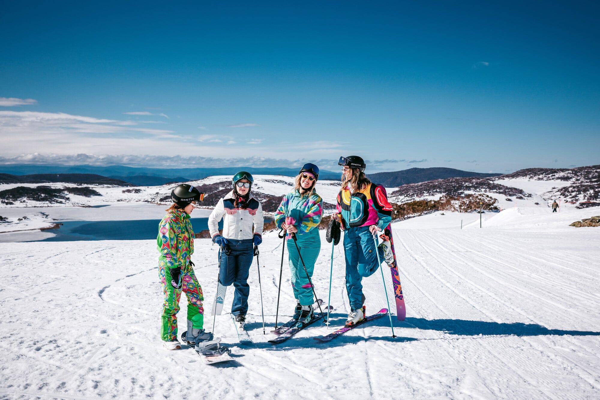 Group of skiers and snowboarders in vintage snow wear at Falls Creek in the snow with mountain and lake views behind them.
