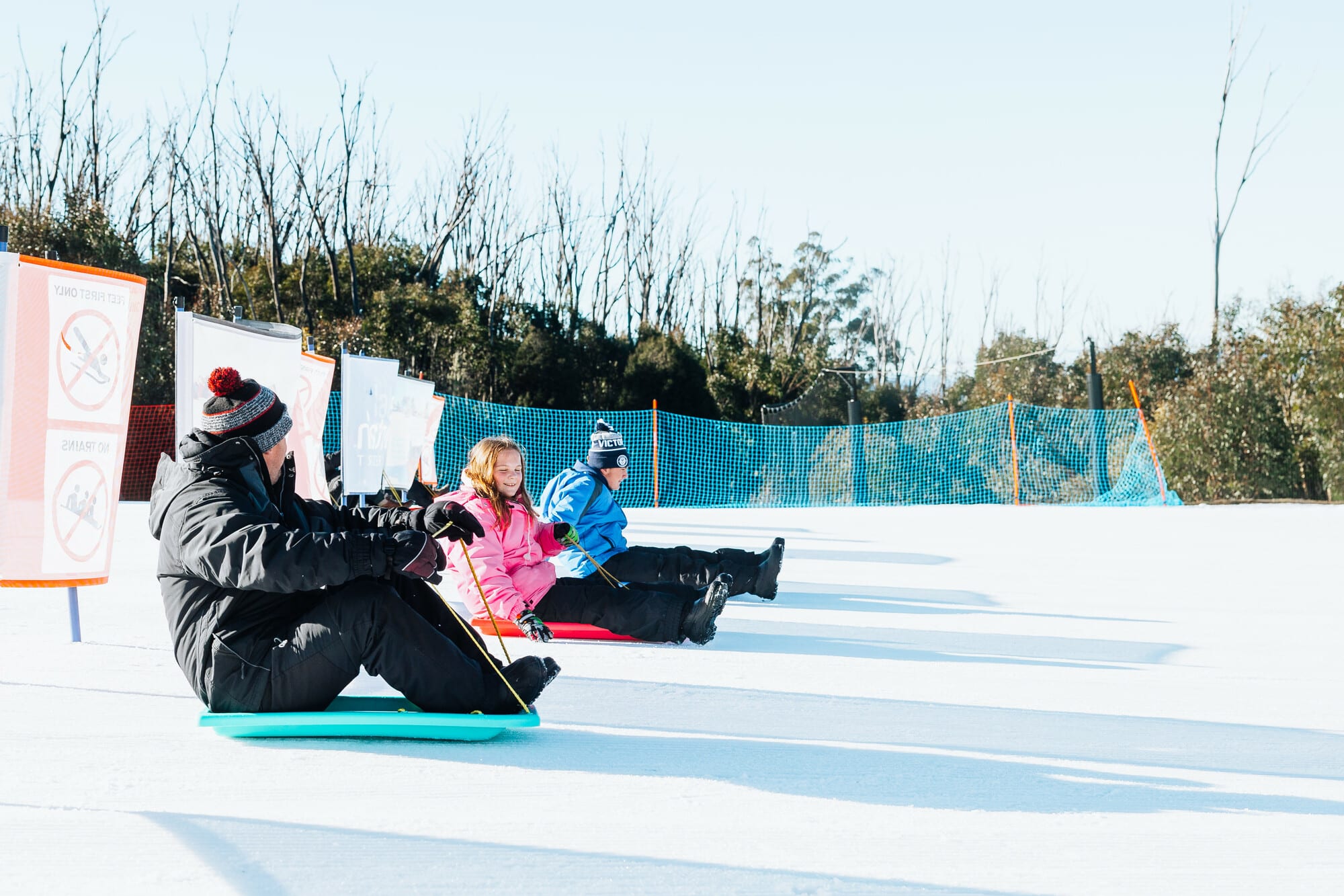 An adult and two children sitting on toboggans at Lake Mountain with blue skies and alpine trees in the background.